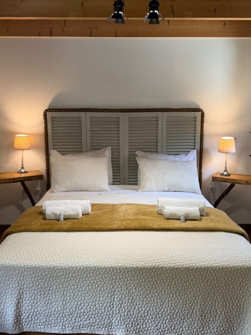 Quarto aconchegante no Refúgio do Nevoeiro com cabeceira veneziana, roupa de cama branca, acabamento dourado, dois abajures e almofadas enroladas