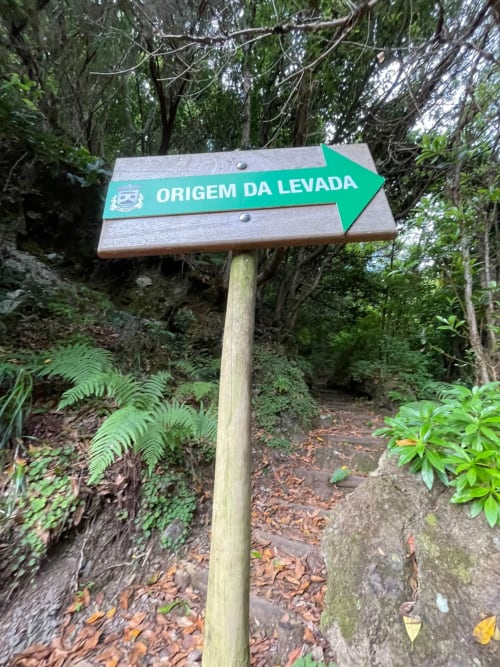 Placa direcional verde apontando para a direita com o texto 'Origem da Levada' na trilha da floresta perto do Refúgio do Nevoeiro, São Vicente
