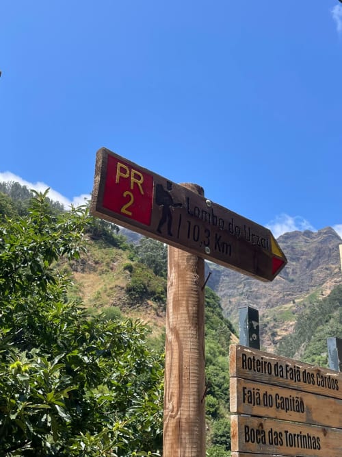 Placa de trilha da PR2 no Refúgio do Nevoeiro em São Vicente, Portugal, rodeada por vegetação verde exuberante e terreno íngreme