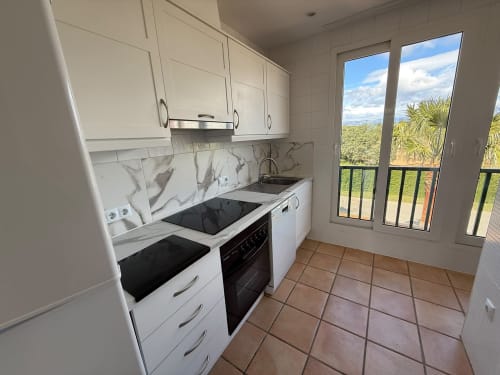 Cocina moderna con gabinetes blancos, backsplash de mármol y balcón con vistas a los jardines del campo de golf en el apartamento Sant Jordi