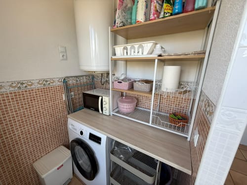 Compacta sala de lavandería en apartamento Sant Jordi con lavadora blanca, microondas, encimera de madera y estantería blanca
