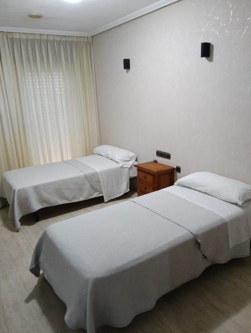 Habitación triple del Mesón Hotel El 21 en Níjar con ropa de cama blanca, mesita de noche de madera, ventana con cortinas y lámpara de pared