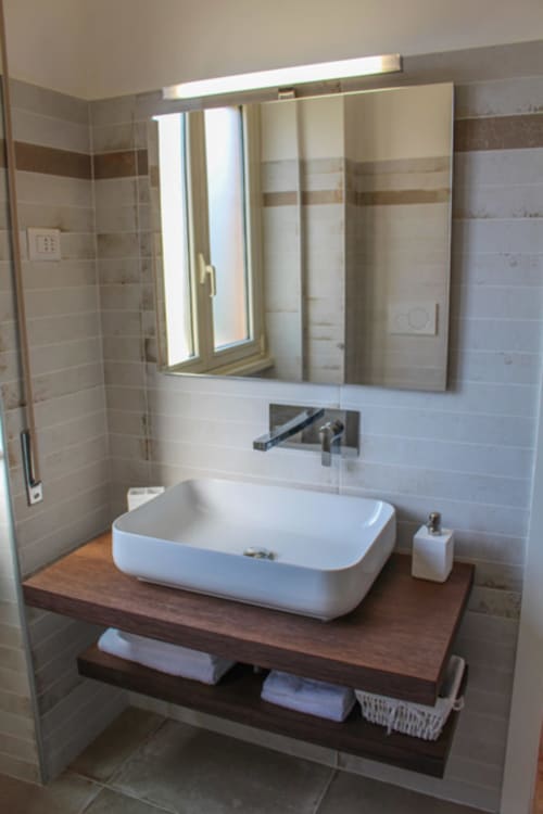 Bagno moderno con lavabo bianco su mobile in legno, rubinetto a parete, specchio illuminato e piastrelle beige