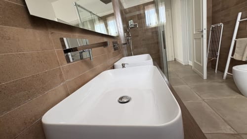 Bagno moderno con due lavabi bianchi, pareti in piastrelle marroni e cabina doccia in vetro presso Maison Tizi Lovely Suites Roma