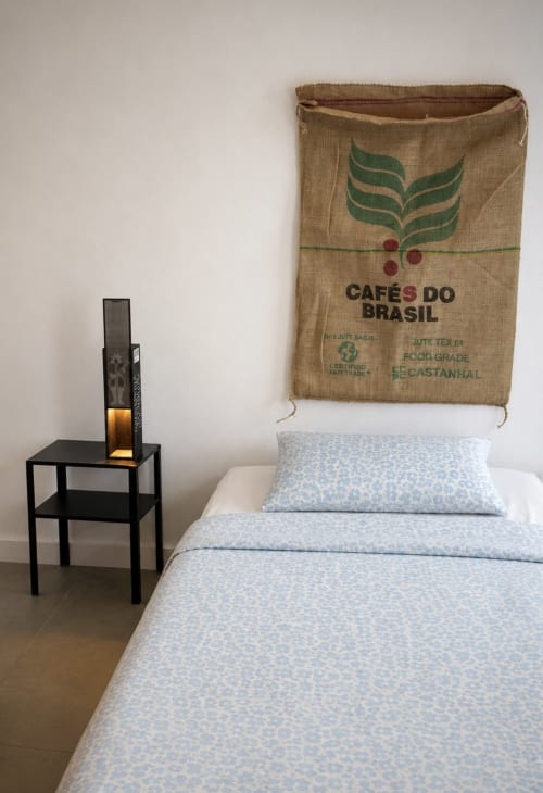 Quarto em Villas Simone Porto Santo com roupa de cama azul, criado-mudo preto e parede decorada com saco de café Cafés do Brasil