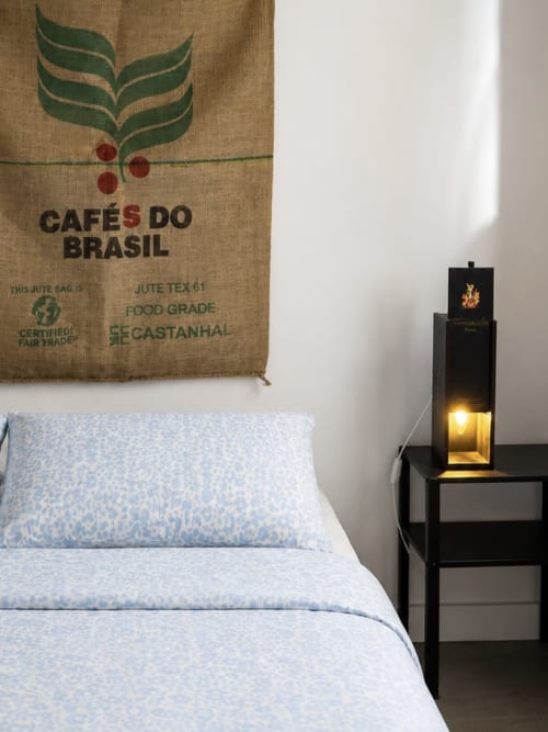Quarto na Villas Simone Porto Santo com parede de arte em saco de café, roupa de cama azul claro e luminária preta na criada