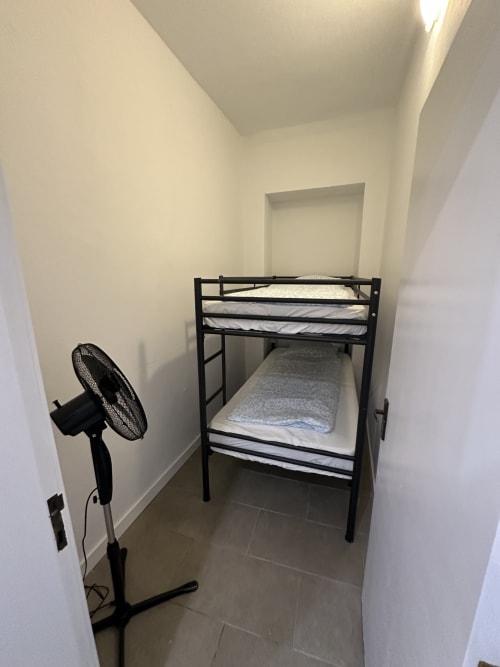 Quarto compacto com beliche de metal preto, roupas de cama brancas e ventilador de pé na Villas Simone Porto Santo