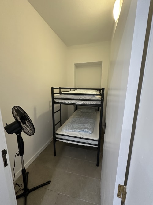 Quarto compacto com beliche de ferro preto, colchões cinzas, ventilador de pé e paredes brancas em Villas Simone Porto Santo