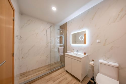 Modern bathroom at Michaels Garden apartment with marble walls, illuminated mirror, white vanity, walk-in shower, and wooden