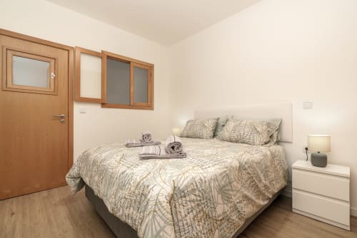 Bright bedroom with patterned bedding, wooden door, windows, white nightstand with lamp, and hardwood floors at Michaels