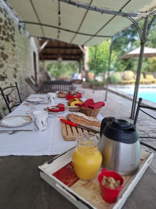 Mesa de desayuno al aire libre en Les Gravets B&B con pan recién hecho, zumo y servilletas rojas bajo pérgola con vistas a la piscina