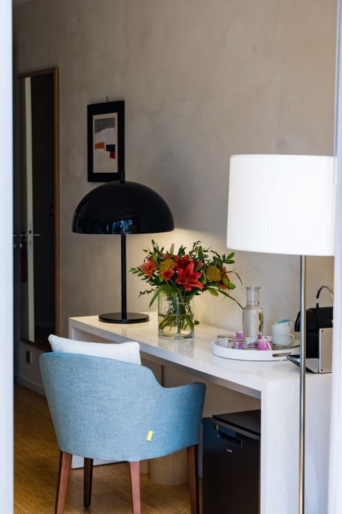 Espace de travail dans une chambre d'hôtel moderne avec chaise bleue, bureau blanc, lampe dome noire et fleurs fraîches au Domaine de la Peyrine