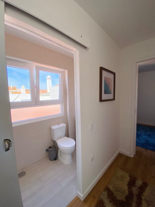 Casa de banho moderna com sanita, janela com vista para os telhados de Peniche, pavimento em azulejo branco no apartamento Casa das 7 Janelas.