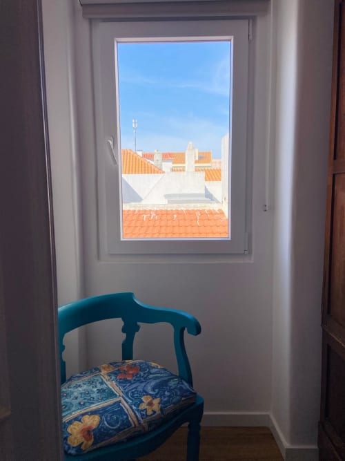 Cadeira verde-azulada com almofada estampada junto à janela, com vista para telhados de terracota em Peniche, Casa das 7 Janelas