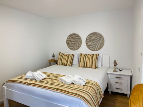 Quarto luminoso na Casa das 7 Janelas em Peniche com cama de listras, travesseiros brancos, arte circular na parede e criado
