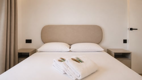 Dormitorio moderno en Holidays Costa Brava con cabecero tapizado beige, ropa de cama blanca y toallas dobladas