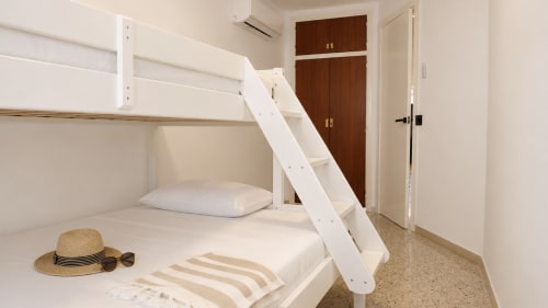 Cama litera moderna con estructura blanca y almohadas a rayas en apartamento Costa Brava, armario de madera y accesorios de playa