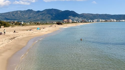 Playa de arena en Empuriabrava con agua turquesa cristalina, montañas y edificios de la localidad costera al fondo