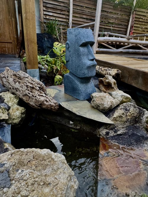 Statue moaï dans un jardin tropical avec bassin à la résidence Tiki Kaz Saint François, décorée de rochers et éléments en bois