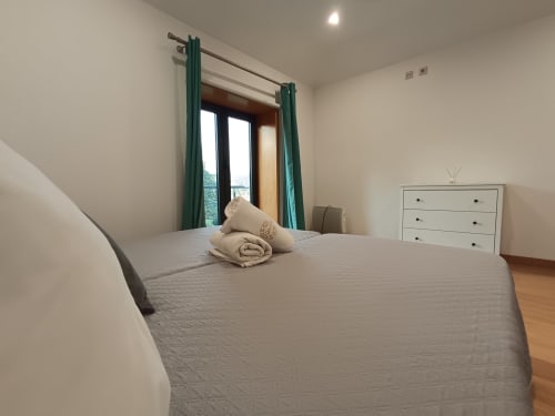 Quarto luminoso na Quinta d'Areda com cama queen, cortinas verdes, vista para o lago, cômoda branca, piso de madeira e comodidades modernas
