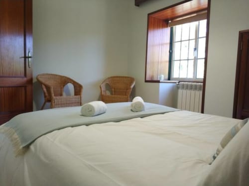 Quarto espaçoso da Quinta d'Areda com roupa de cama branca, toalhas enroladas, cadeiras de vime e luz natural das grandes janelas