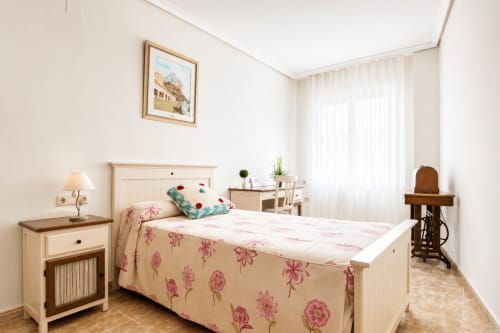 Dormitorio luminoso en apartamento Riurau Home Quintanes, Calp, con ropa de cama floral, muebles de madera y gran ventana con luz natural