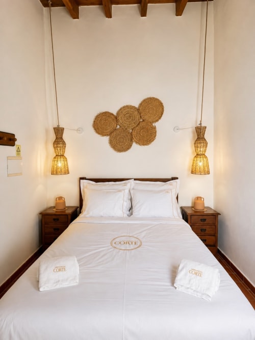 Quarto luminoso na Herdade da Côrte com cama queen, cestos de palha na parede, luminárias pendentes, criados de madeira e roupa de cama branca