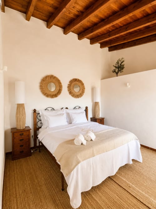 Quarto rústico com vigas de madeira, roupa de cama branca, espelhos tecidos e piso em terracota na Herdade da Côrte, Tavira
