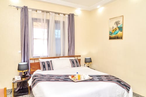 Chambre standard de l'Hôtel Yakro à Yamoussoukro avec lit en bois, oreillers rayés, lampes modernes et fenêtre aux volets pourpres
