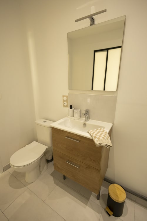 Salle de bains moderne avec vasque blanche, miroir lumineux, toilettes et poubelle noire au gîte Accolade Chez Ninon et Lou