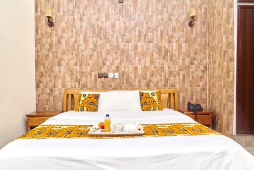 Suite de l'Hôtel Yakro à Yamoussoukro avec mur d'accent en carreaux de bois, literie blanche, coussins dorés et plateau d'équipements