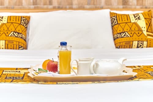 Petit-déjeuner en suite à l'Hôtel Yakro avec jus, pomme, théière blanche et coussins à motifs jaunes sur le lit