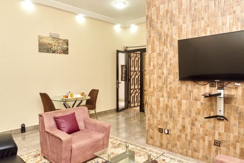 Suite moderne à l'Hôtel Yakro avec fauteuil rose, téléviseur écran plat, table à manger et mur de briques à Yamoussoukro