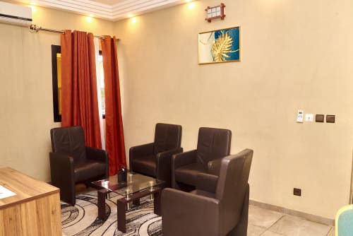 Suite Yakro de l'hôtel à Yamoussoukro avec sièges en cuir marron, table en verre, rideaux orange et accès à un balcon avec vue sur la ville