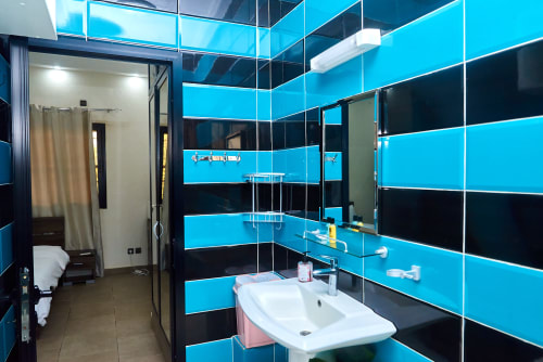 Salle de bain moderne de l'Hôtel Yakro avec carrelage bleu vif et noir, lavabo blanc, miroir et douche à Yamoussoukro