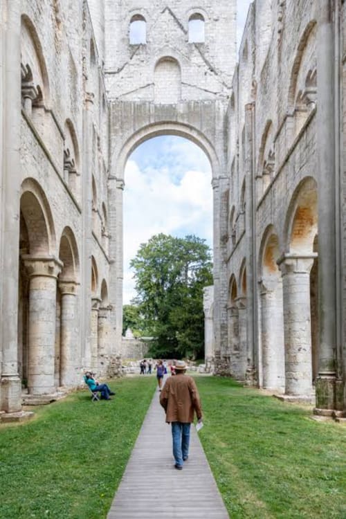 Ruines abbatiale en pierre ancienne avec colonnes voûtées, pelouse verdoyante et visiteurs explorant le site historique du Héron, France