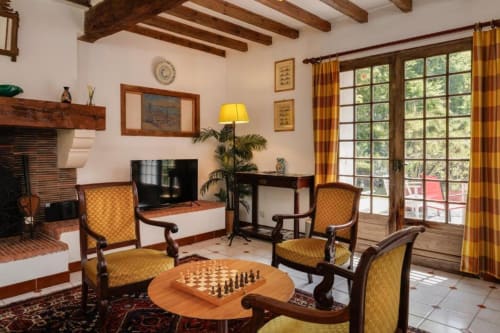 Salon rustique du gîte Les Petits Toits avec cheminée, poutres en bois, table d'échecs et vue sur le jardin