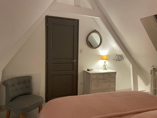 Chambre cosy mansardée aux Gîtes du Grand Saule à Tardinghen avec plafonds pentes, fauteuil gris et commode en bois.