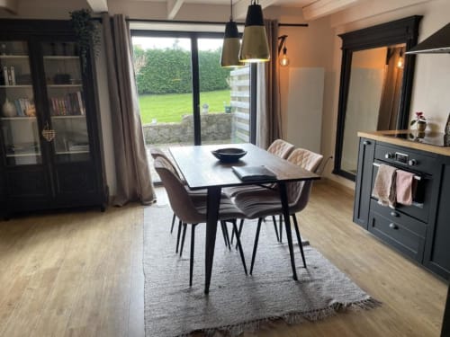 Salle à manger de gîte moderne avec vue sur le jardin, table noire, chaises beige, cuisine et grandes fenêtres aux gîtes du grand