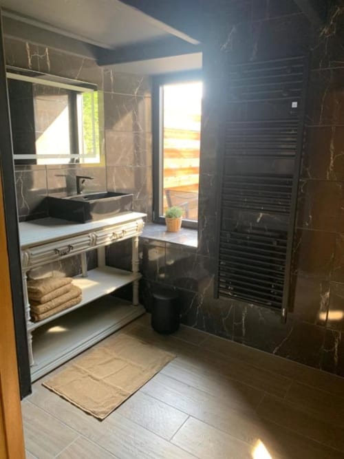 Salle de bains moderne aux murs en marbre noir, vasque avec robinetterie noire, douche spacieuse et porte donnant sur le jardin aux Gîtes du