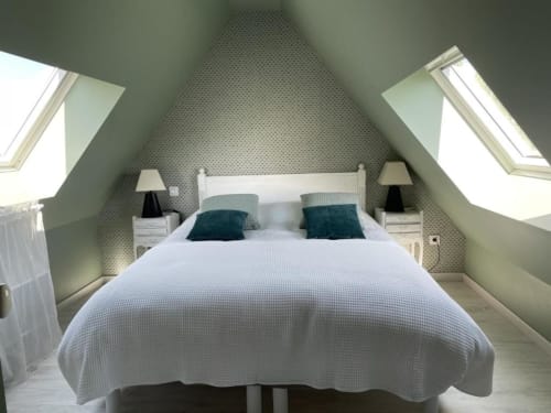 Chambre lumineuse sous les toits avec lit blanc, oreillers bleu-vert, verrières et papier peint à motifs - Les gîtes du grand saule