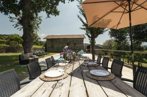 Terraço de refeições ao ar livre da Quinta de Carvalhal com casa de pedra, guarda-sóis e jardins ondulantes em Guimarães