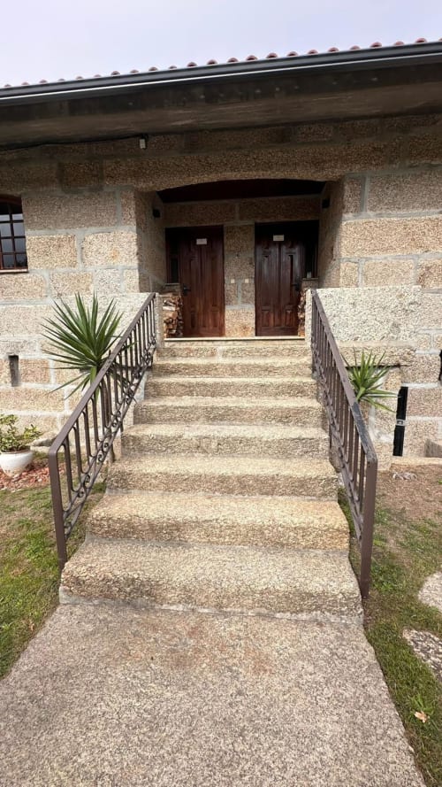Entrada da casa de pedra na Quinta de Carvalhal, Guimarães, com portas de madeira, grades de ferro e plantas do jardim