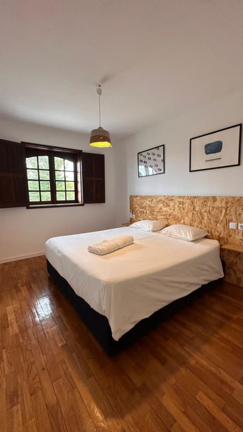 Quarto espaçoso com cama de casal, cabeceira de cortiça, assoalho em madeira, luminária suspenso e janela com vista para jardim na Quinta de Carvalhal