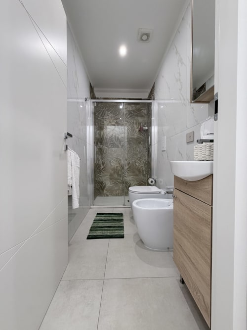 Bagno moderno a Sabò Bologna con piastrelle in marmo bianco, doccia in vetro, bidet e wc bianchi, mobile lavabo in legno e dettagli a righe verdi