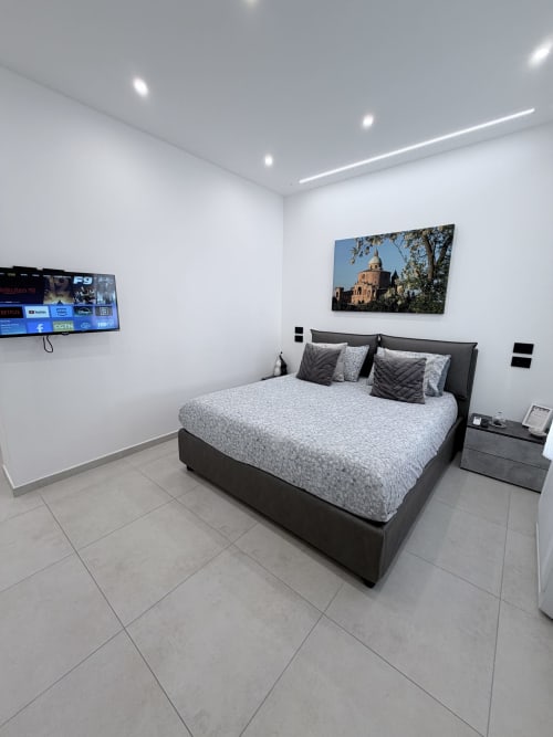 Camera moderna a Sabò Bologna con letto scuro, biancheria grigia, TV a parete e fotografia architettonica sopra la testata