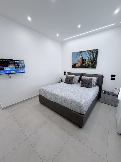 Camera moderna a Sabò Bologna con letto nero, biancheria grigia, TV a parete e opere d'arte della cattedrale