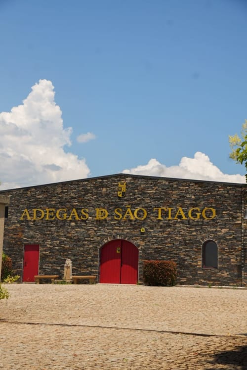 Fachada de pedra da adega d'São Tiago com portas vermelhas em Dominguiso, Portugal sob céu azul