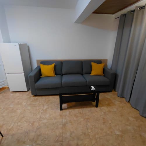 Sala de estar moderna en la casita El Palm-Mar con sofá negro, cojines amarillos, techo de madera y suelo de corcho