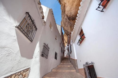 Callejón estrecho de paredes blancas en Setenil de las Bodegas con escalones de piedra, ventanas con rejas de hierro y roca tallada sobresaliente